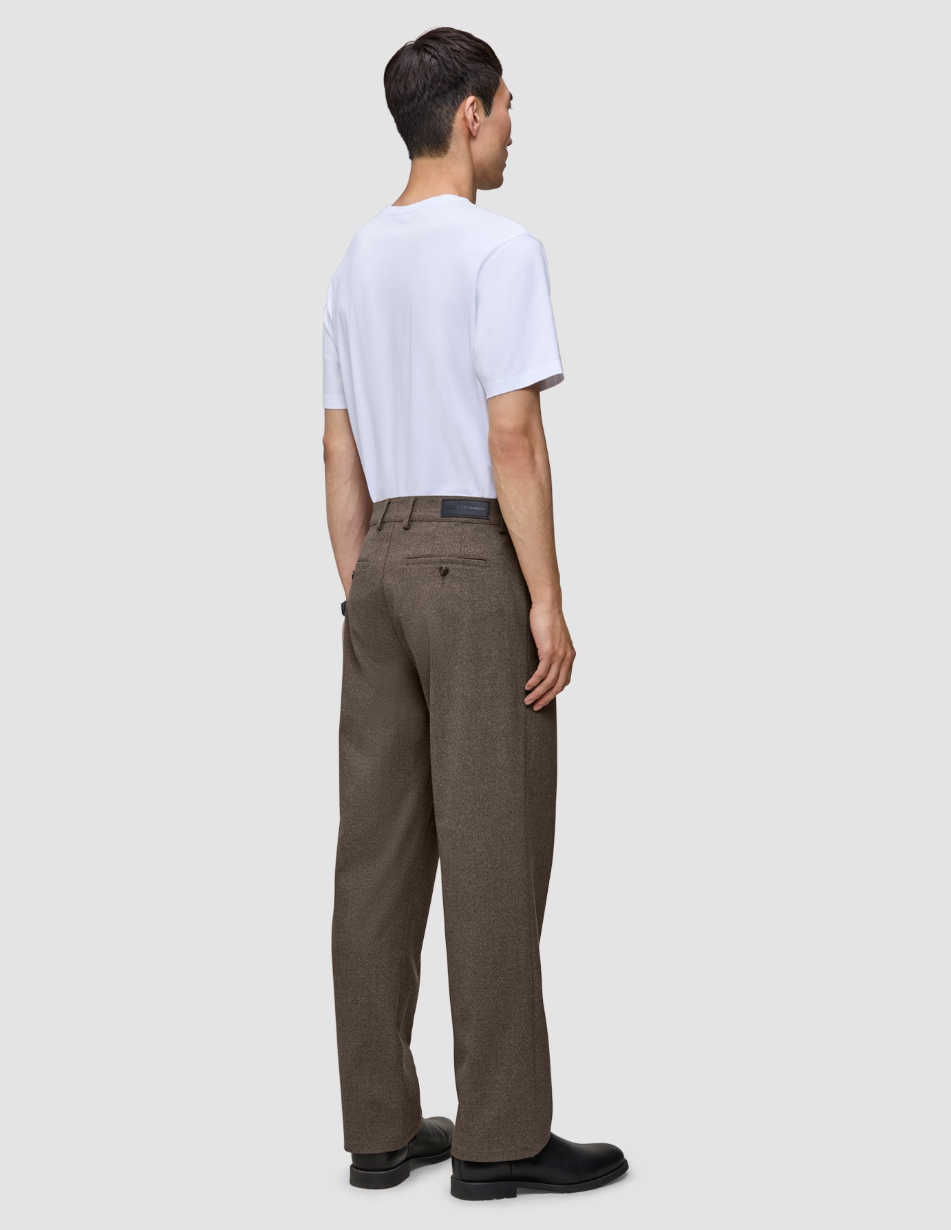 En man sedd bakifrån, klädd i en vit t-shirt och ett par löst sittande byxor i skandinavisk design, Atelier Pants Relaxed Dark Beige.